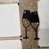 Decorative card with black wine glass cutouts with gold sequins on a patterned background.